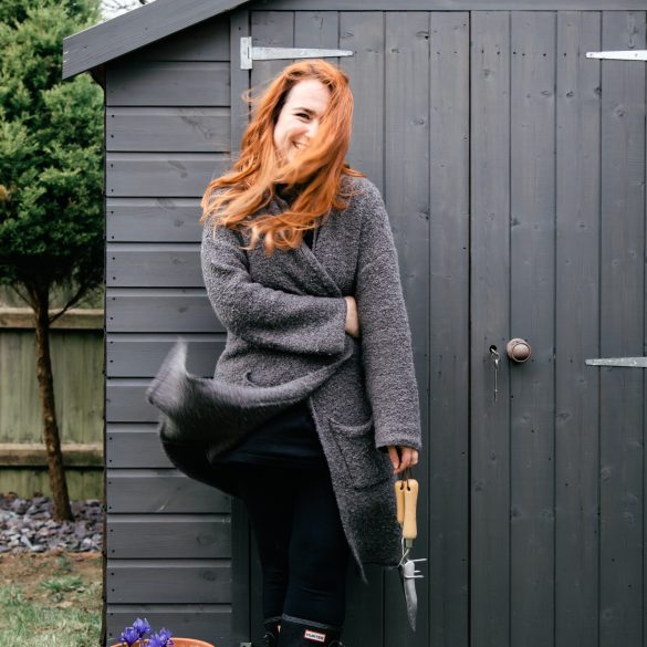 Woman stood in front of grey shed with garden tools in hand and flower pots to the side